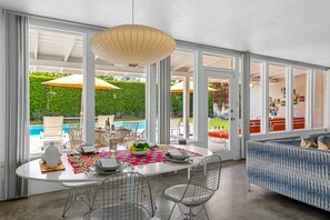 Quarto amplo com mesa de jantar, cadeiras, sofá e janelas grandes que dão para uma piscina e áreas verdes.