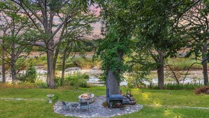 Een rustig buitenzitgedeelte met een vuurkorf, een barbecue en een stenen tafel, omringd door weelderige begroeiing en uitzicht op een rivier.