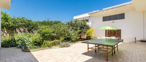 A patio area with a ping pong table, a green umbrella, and a white building with a window and door.