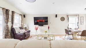 A living room with a leather sofa, a glass coffee table, a dining table with chairs, and a TV mounted on the wall displaying the Netflix logo.