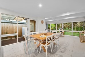 A spacious patio with a wooden table and white chairs, large windows offering a view of the garden, and a wooden fence in the background.
