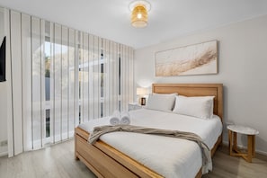 A modern bedroom with a wooden bed, a nightstand, a small table, and a large window with vertical blinds.