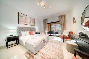 A modern bedroom with a bed, bedside tables, a desk, and a chair. There is a large window with curtains and a view of buildings outside.