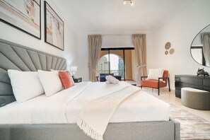 Une chambre à coucher moderne avec un grand lit, une tête de lit grise, une table de chevet, une chaise, une commode et une vue sur la ville par une grande fenêtre.