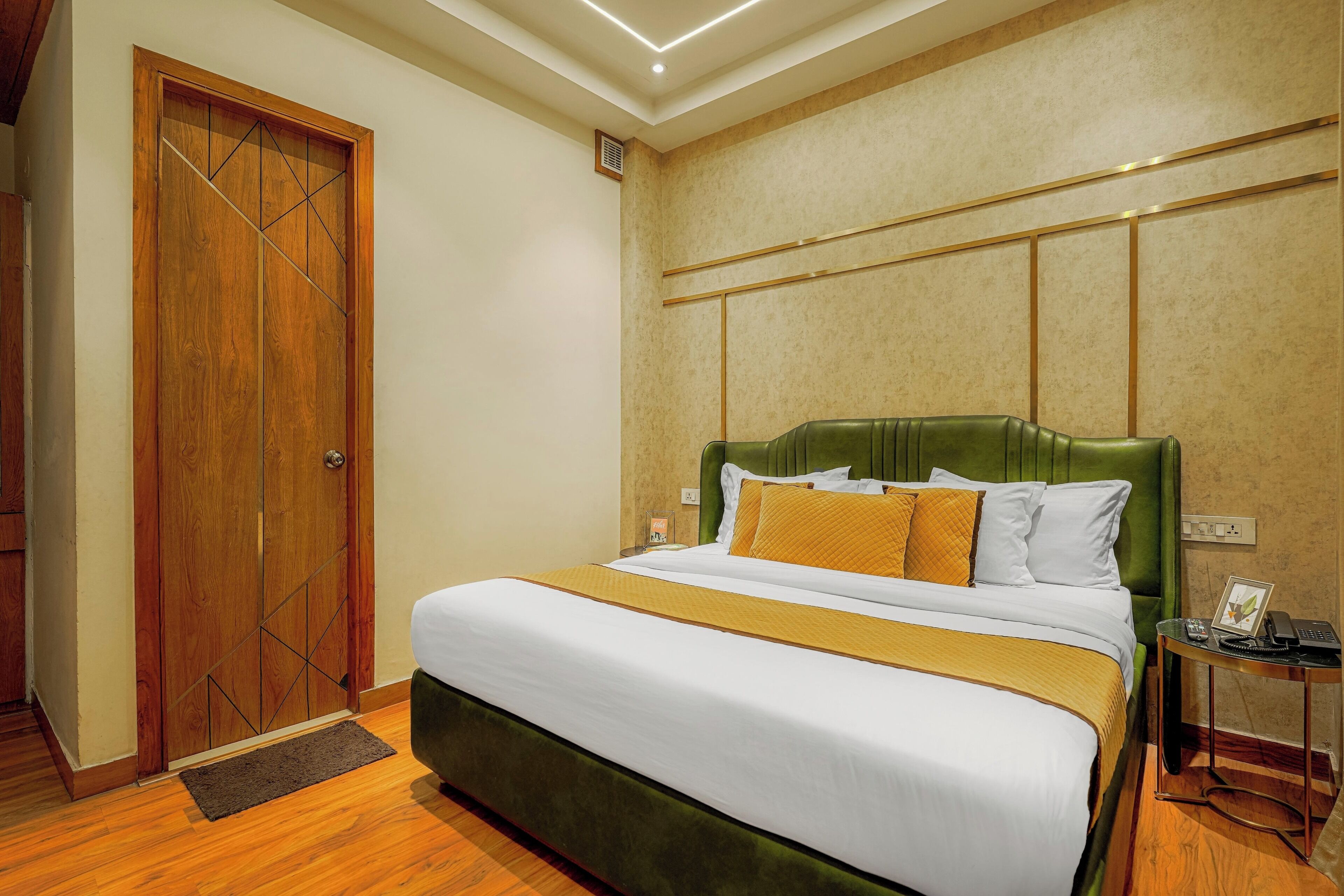 A hotel room with a large bed, a wooden door, a green upholstered headboard, and a bedside table with a telephone and a picture frame.