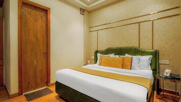 A hotel room with a large bed, a wooden door, a green upholstered headboard, and a bedside table with a telephone and a picture frame.