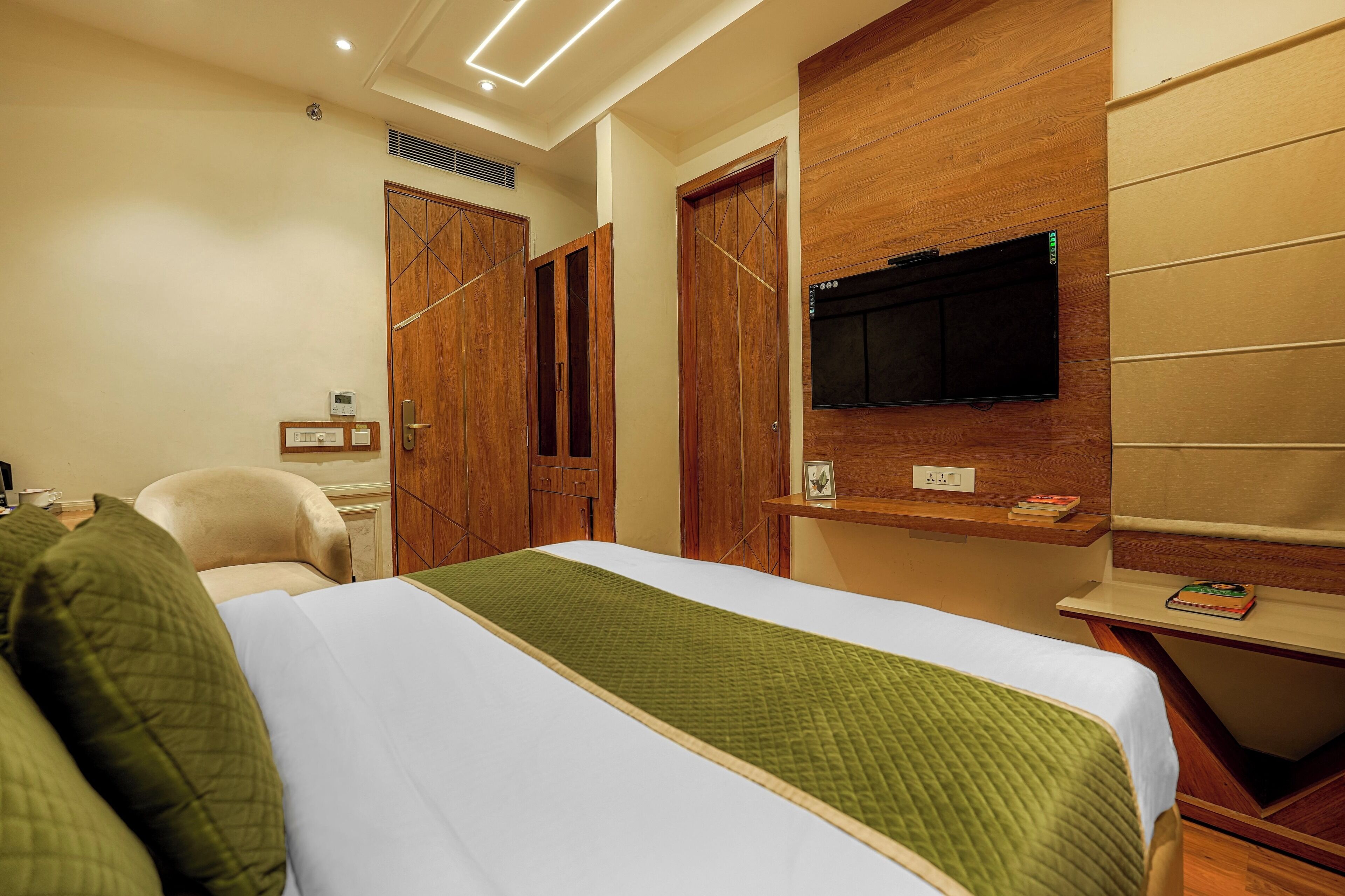 A hotel room with a bed, a TV mounted on the wall, wooden doors, and a small shelf with a book.