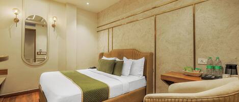 Een hotelkamer met een bed, een stoel, een klein tafeltje met flessen en een mand, en een spiegel aan de muur.