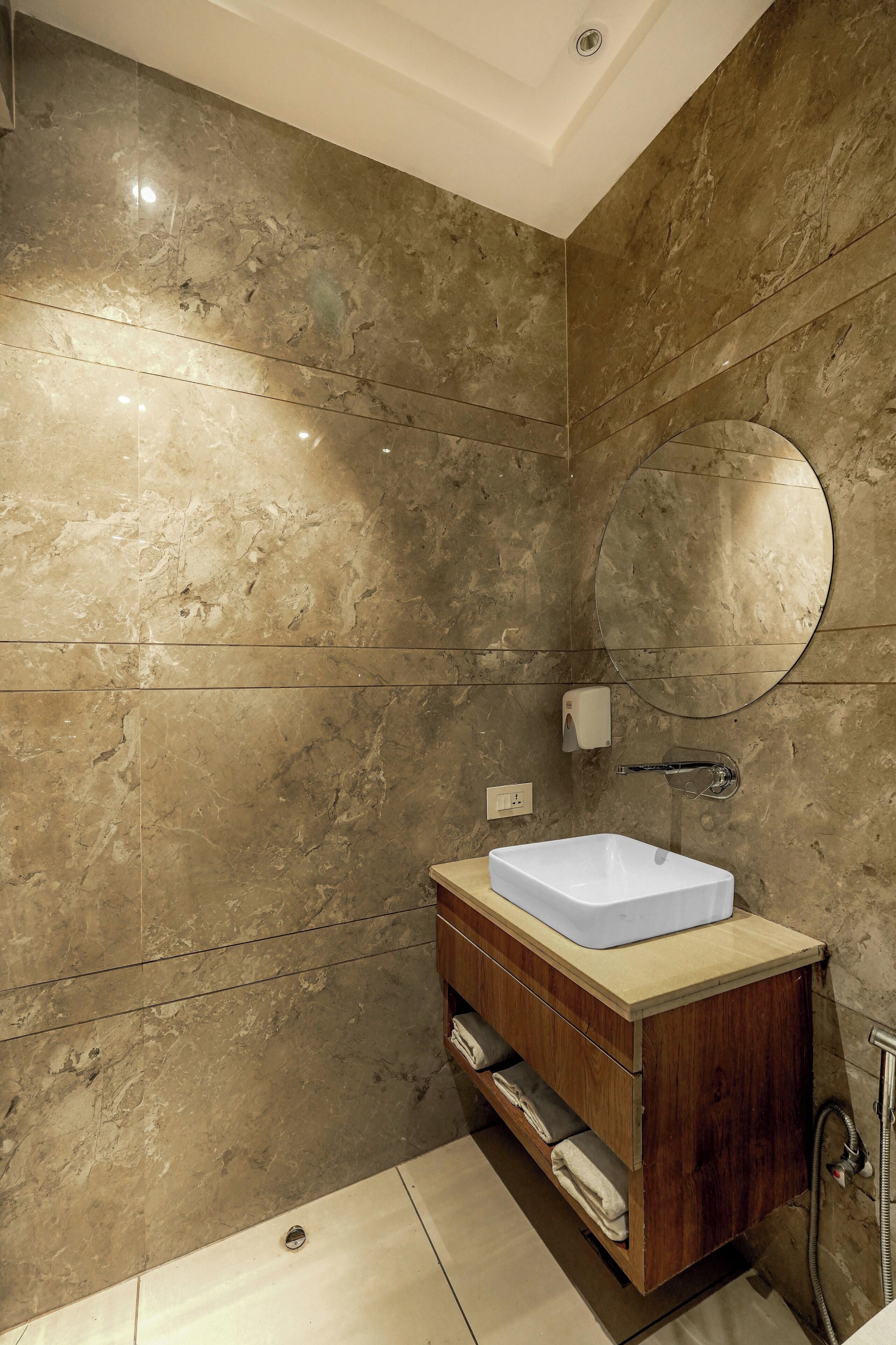 A bathroom with a marble wall, a round mirror, a white sink, and a wooden vanity.