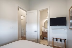 Un dormitorio con cama, televisor montado en la pared, puerta que da a un baño y cabecera de madera.