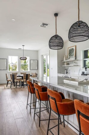 A modern kitchen with a central island, bar stools, pendant lights, and large windows.