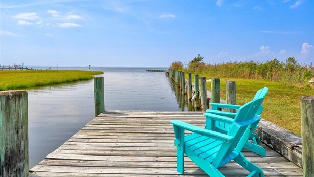 Un muelle de madera que se adentra en aguas tranquilas, con una silla Adirondack turquesa a la derecha.