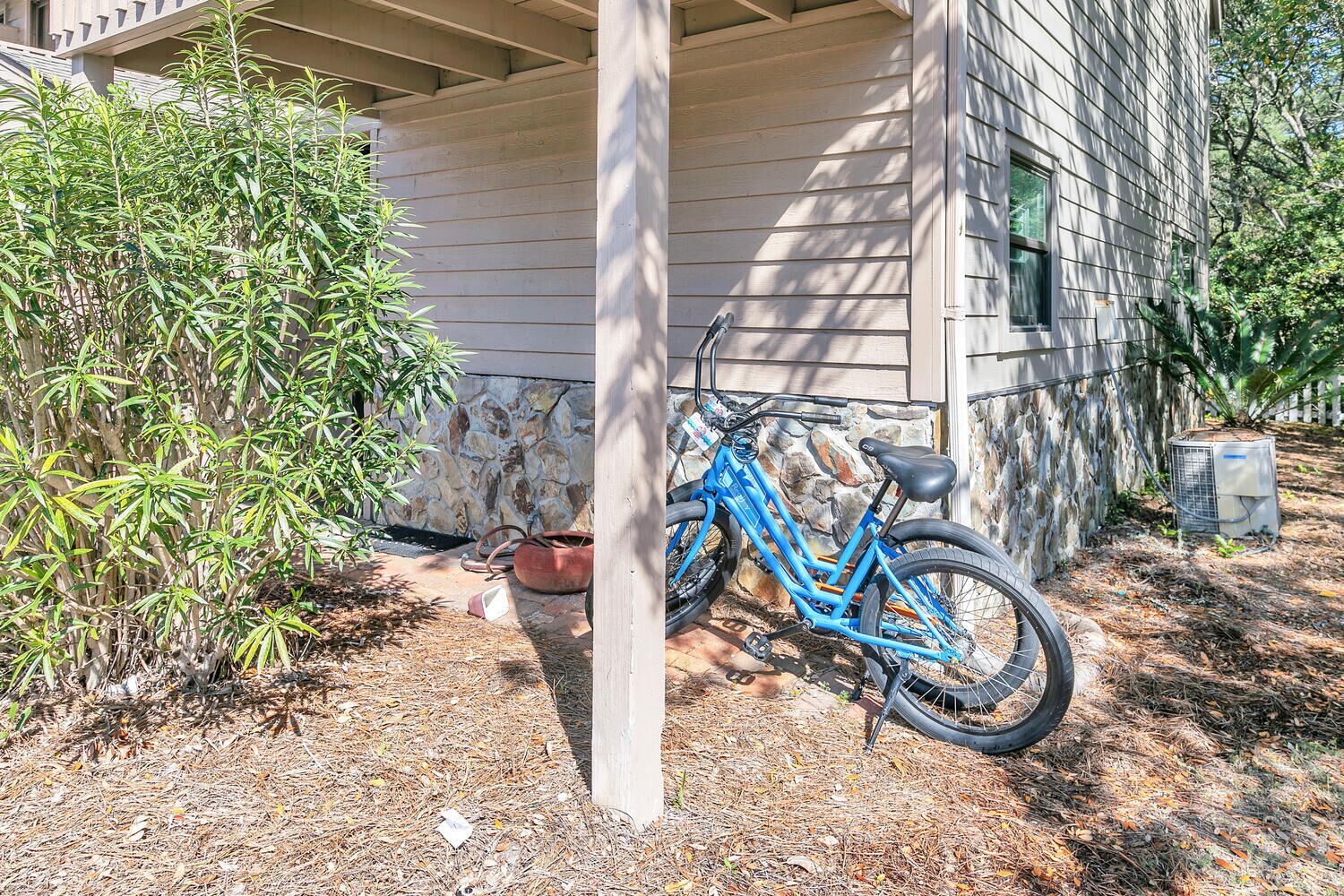 Una bicicleta azul estacionada bajo una estructura de madera con un muro de piedra y una planta en maceta cerca.