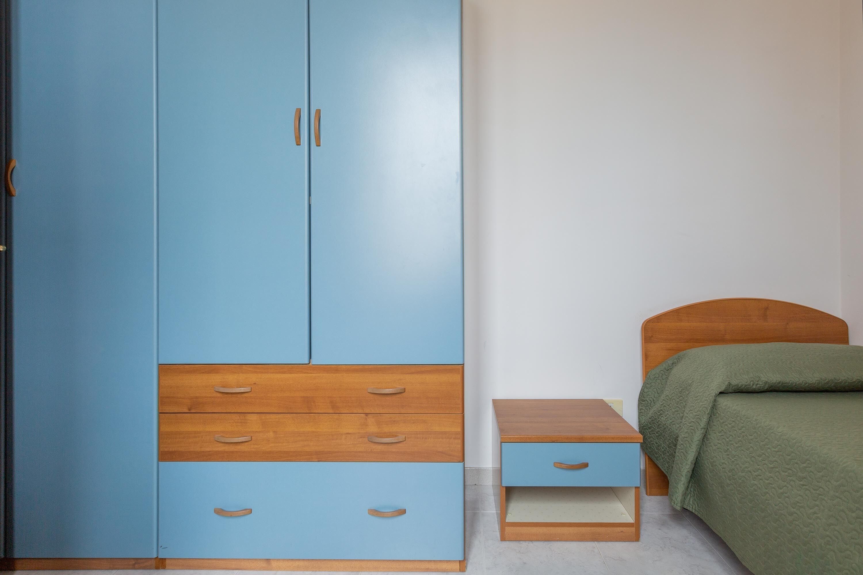 A bedroom with a blue wardrobe, a wooden bedside table, a green bedspread, and a wooden headboard.