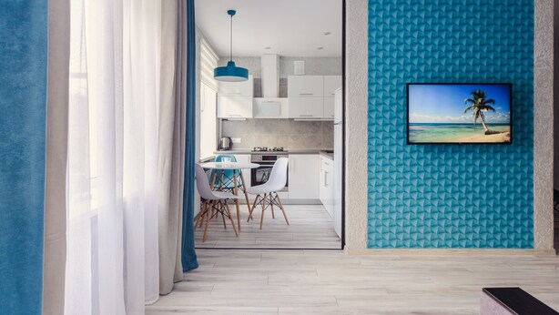 A modern kitchen with white cabinets, a dining area with a round table and chairs, and a TV mounted on a textured blue wall. - Cozy apartment on Vera Khoruzhey street (Minsk)
