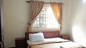 A bedroom with a bed, a nightstand, a dresser, and a window with curtains. - Sycamore Royals Hotel Jahi (Abuja)