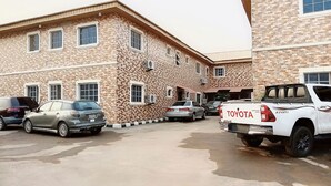 A building with a brick facade, multiple windows, and parked cars including a white Toyota pickup truck. - Sycamore Royals Hotel Jahi (Abuja)