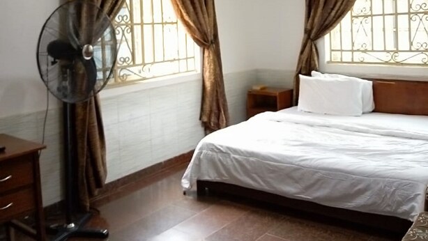 A hotel room with a bed, a bedside table, a fan, and curtains. - Sycamore Royals Hotel Jahi (Abuja)