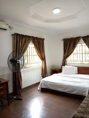 Une chambre d’hôtel avec un lit, une table de chevet, un ventilateur et des rideaux.