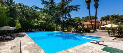 A swimming pool with clear blue water, surrounded by stone tiles and shaded by trees and umbrellas.
