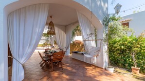 Eine überdachte Terrasse mit Tisch, Stühlen, weißen Vorhängen und Blumentöpfen.