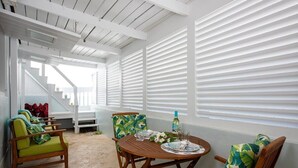 Un área de estar al aire libre con techo, muebles de madera y contraventanas blancas.