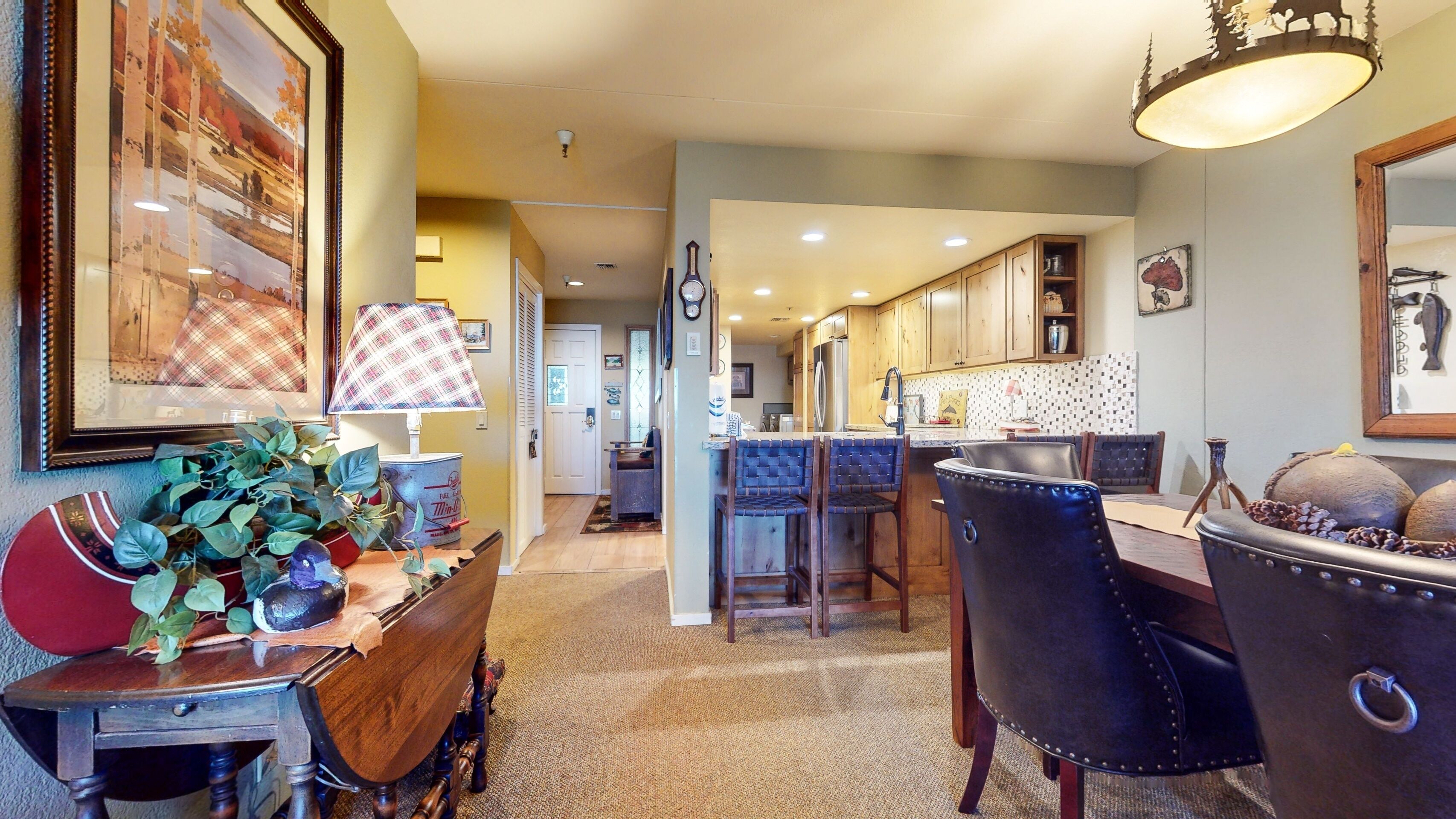 A dining area with a wooden table, chairs, and a lamp. There is a kitchen in the background with wooden cabinets and a tiled backsplash.