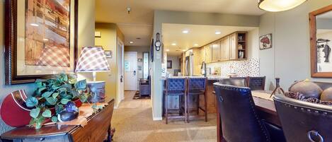A dining area with a wooden table, chairs, and a lamp. There is a kitchen in the background with wooden cabinets and a tiled backsplash.
