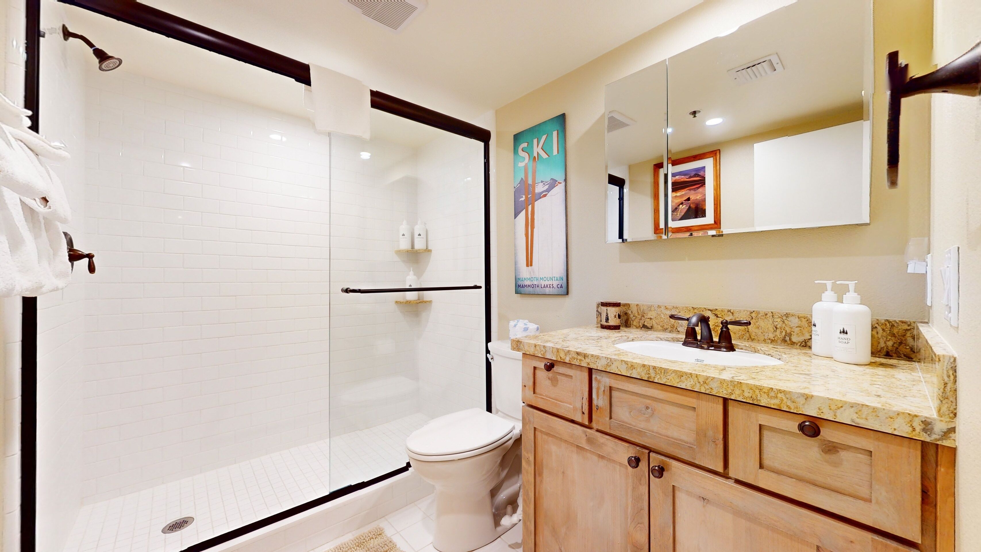 A bathroom with a glass shower enclosure, a toilet, a sink with a marble countertop, wooden cabinets, and a large mirror.