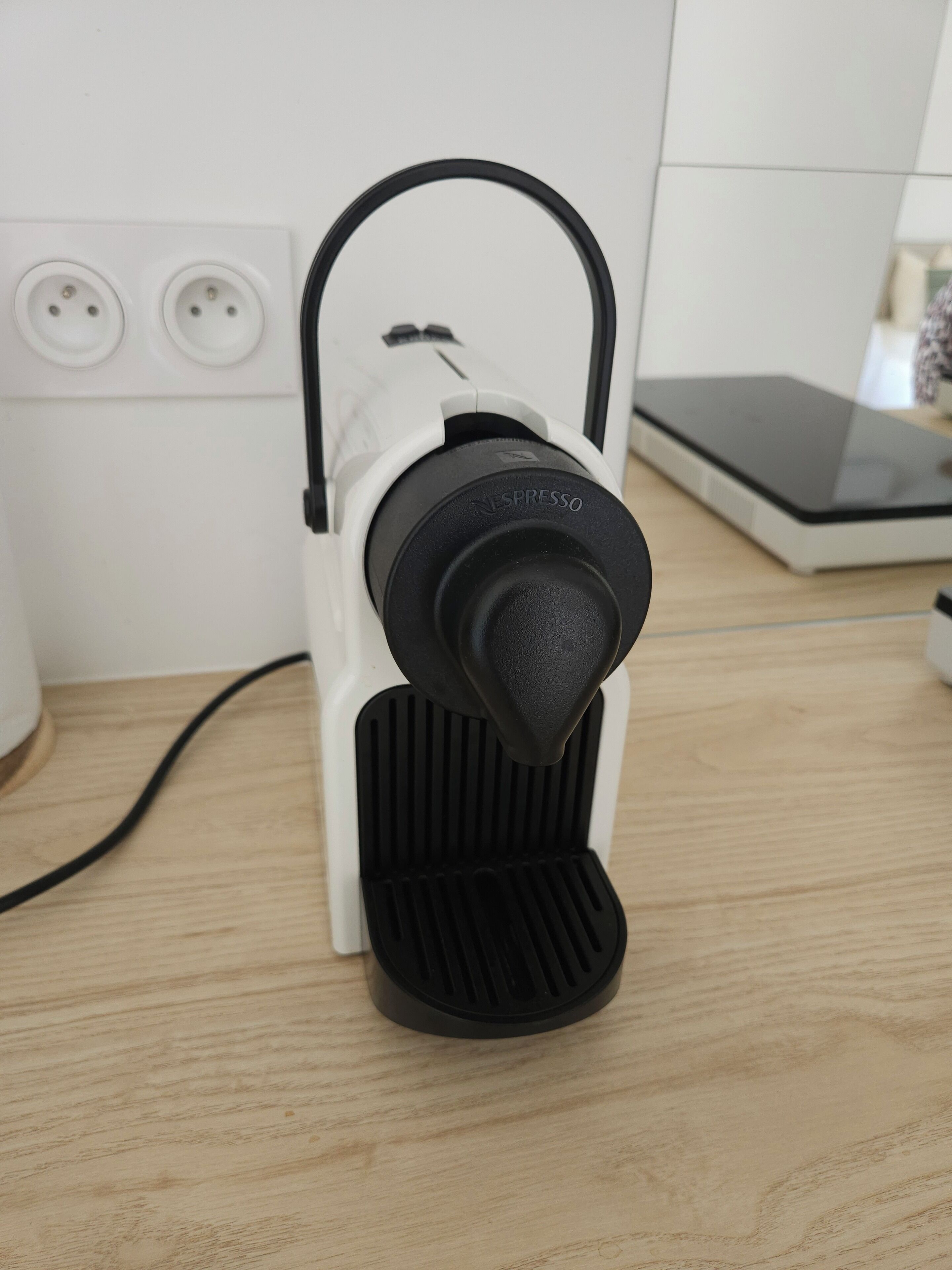 Une machine à café Nespresso posée sur une surface en bois, avec une prise de courant en arrière-plan.
