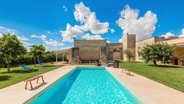 A rectangular swimming pool with a clear blue water surface, surrounded by a paved deck. There are two wooden benches and two blue lounge chairs on the deck. The pool area is adjacent to a building with stone walls and a flat roof. - 3625 Villa Queen by Perle di Puglia (Lecce)