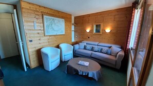 Ein gemütliches Zimmer mit einer Holzwand, einem grauen Sofa, zwei weißen Sesseln und einem kleinen runden Tisch mit einer Zeitschrift.