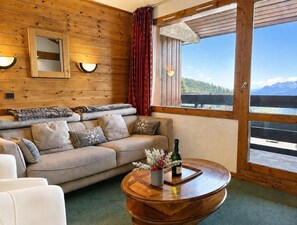 Ein gemütliches Zimmer mit einer Holzwand, einer Couch mit Kissen, einem runden Holztisch mit einer Flasche und Blumen sowie einem großen Fenster mit Bergblick.
