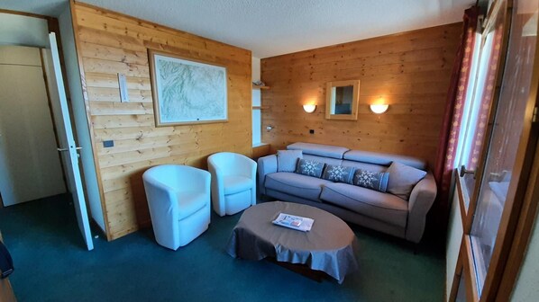 Una habitación acogedora con pared de madera, un sofá gris, dos sillones, una mesita redonda y una puerta.