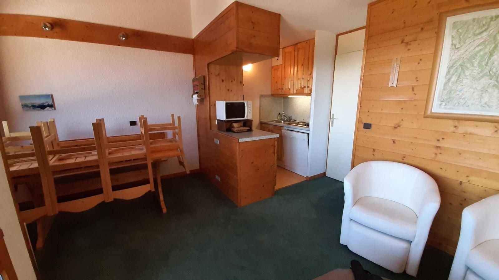 A compact room with a dining table, chairs, a small kitchenette, and a white armchair.
