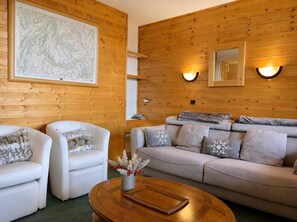 Una habitación con paneles de madera, dos sillones blancos, un sofá gris y una mesa de centro de madera.