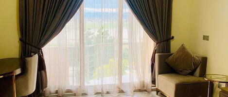 A hotel room with a bed, a chair, a desk, and a view of greenery through sheer curtains.