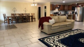 A living room with a beige sofa, a patterned rug, a dining area with wooden chairs, and a kitchen with wooden cabinets and stainless steel appliances.