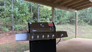 A portable gas grill with a digital control panel, four burners, and a side burner, set up on a concrete patio with a forested background.