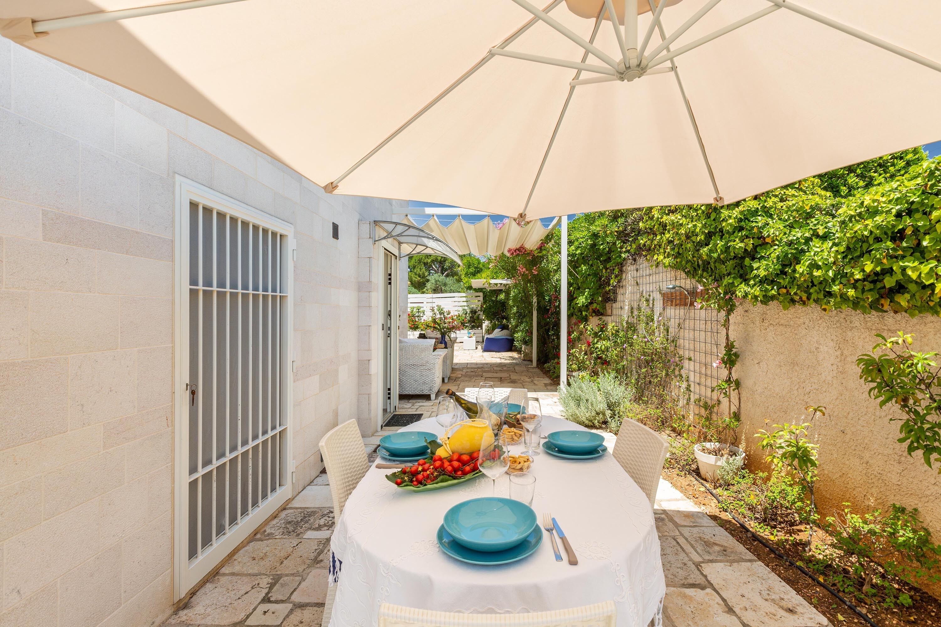 A covered outdoor dining area with a table set for four, surrounded by a garden and a white wall.