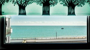 Vista do mar por uma janela com grade, um barco ao longe e um mural de árvore na parede.