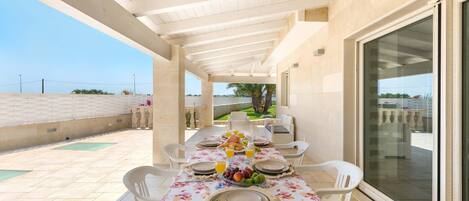 A covered outdoor dining area with a table set for four, surrounded by white chairs, and a glass door leading to an outdoor space.