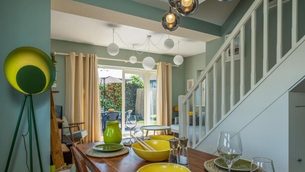 A dining area with a wooden table set for two, a green lamp, and a view of a garden through a window. - Garden apartment with parking in Rye (Rye)