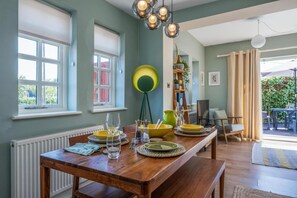 Un coin repas avec une table en bois dressée pour quatre personnes, une fenêtre avec des stores et un luminaire suspendu.