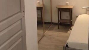 Un couloir étroit avec un plancher en bois, une porte blanche sur la gauche, un miroir sur la droite et une petite table avec une lampe.