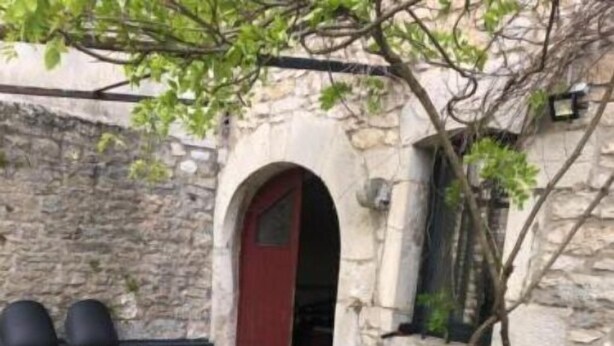 Une table à plateau en verre, entourée de chaises en osier, dans un espace patio extérieur avec des murs en pierre et une porte rouge.