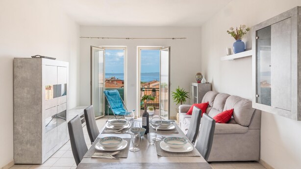 Un coin repas avec une table dressée pour quatre personnes, un canapé gris agrémenté de coussins rouges et une vue sur la mer par de grandes fenêtres.