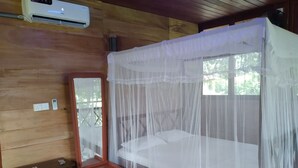 A wooden cabin room with a bed under a mosquito net, a small desk with a mirror, and an air conditioning unit. - Green Herbal Ayurvedic Eco-Chalets  (Unawatuna)