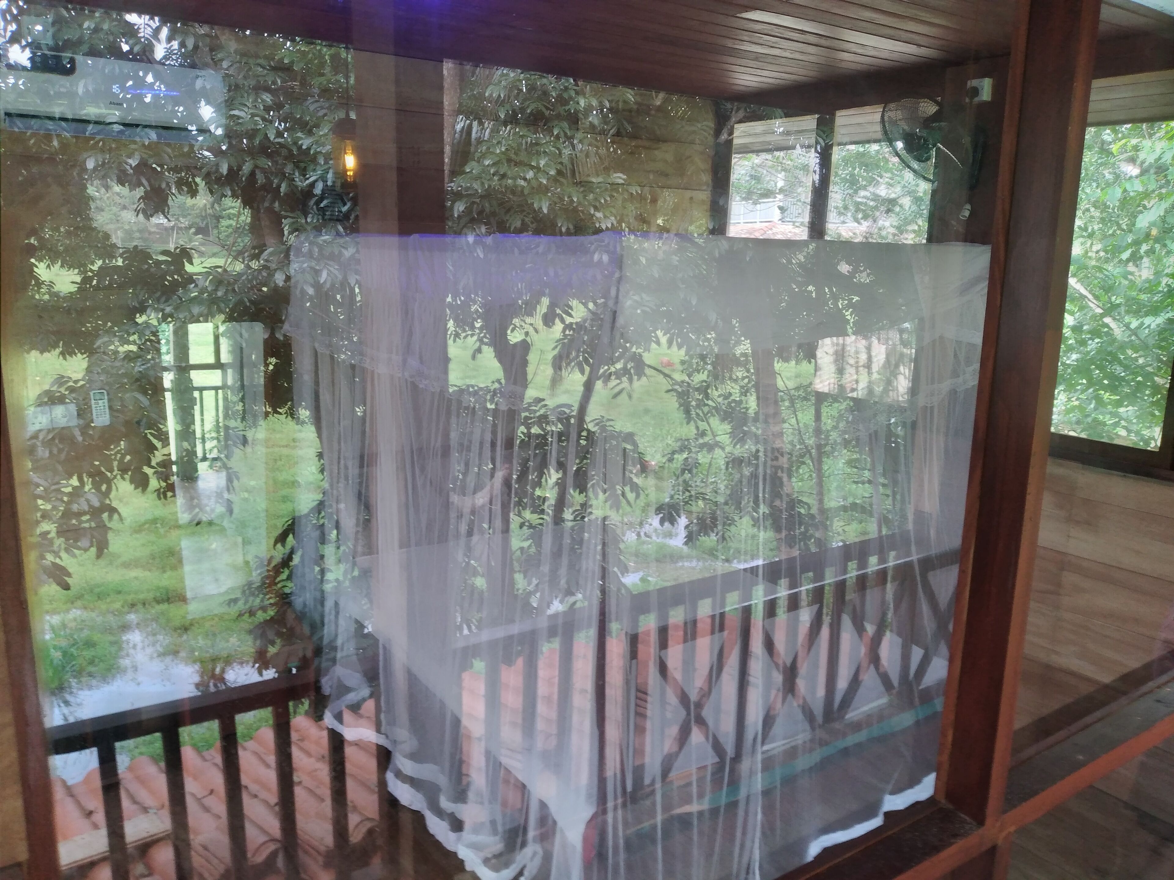 Ein Zimmer mit Holzbalkon, Glastür mit weißem Vorhang und Blick auf einen Garten mit Bäumen und einem kleinen Wasserspiel.