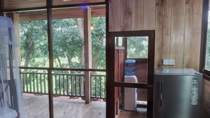 A room with a wooden floor, a large sliding glass door leading to an outdoor area with a wooden deck and a view of trees. There is a water dispenser and a refrigerator on the deck. - Green Herbal Ayurvedic Eco-Chalets  (Unawatuna)